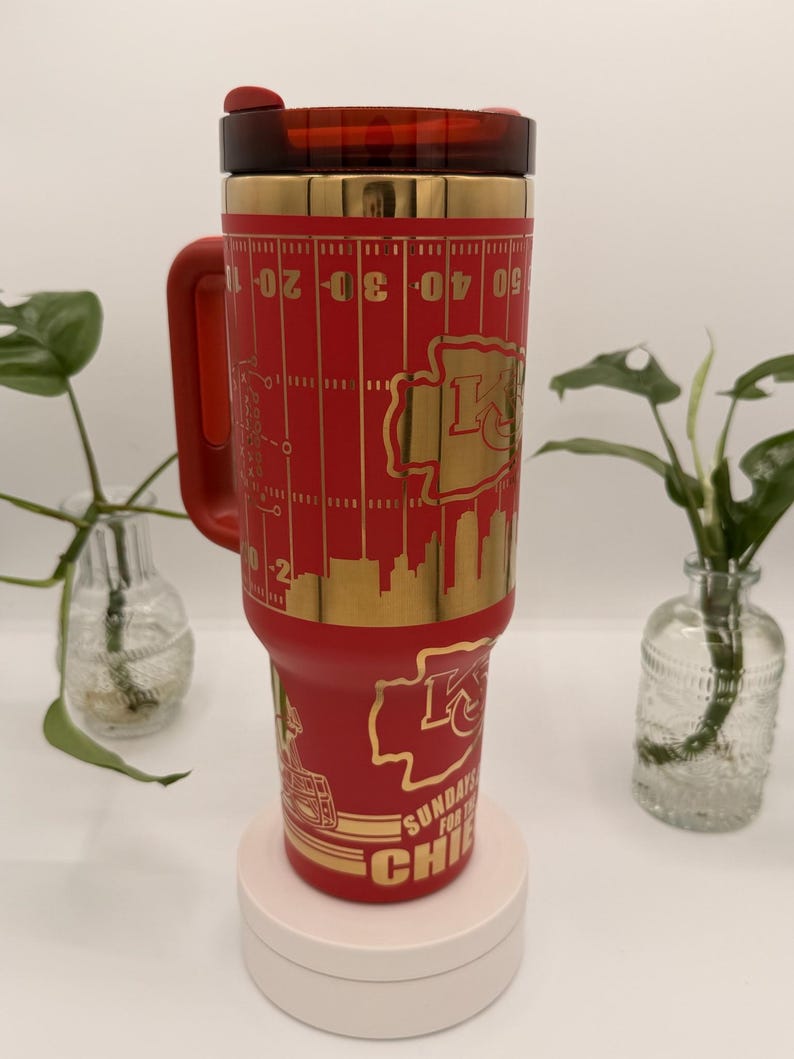 Puede incluir: Un vaso t&eacute;rmico rojo con un dise&ntilde;o dorado que presenta un campo de f&uacute;tbol americano, el logotipo de los Kansas City Chiefs y el texto "SUNDAYS FOR THE CHIEFS". El vaso tiene un asa y una tapa roja.