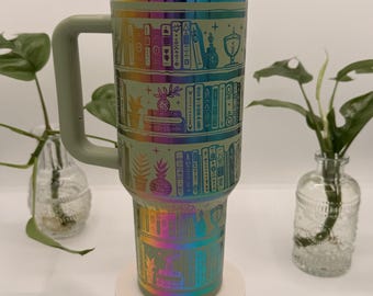 May include: A large, insulated tumbler with a handle, featuring a wraparound design of bookshelves with books, plants, and decorative elements. The tumbler has a green lid and handle, and a rainbow iridescent finish.
