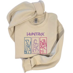 May include: Cream-colored sweatshirt with the word "HUNTR/X" in purple and three outlined figures in boxes. The figures are in blue, purple, and red. The sweatshirt is folded with the hood visible.