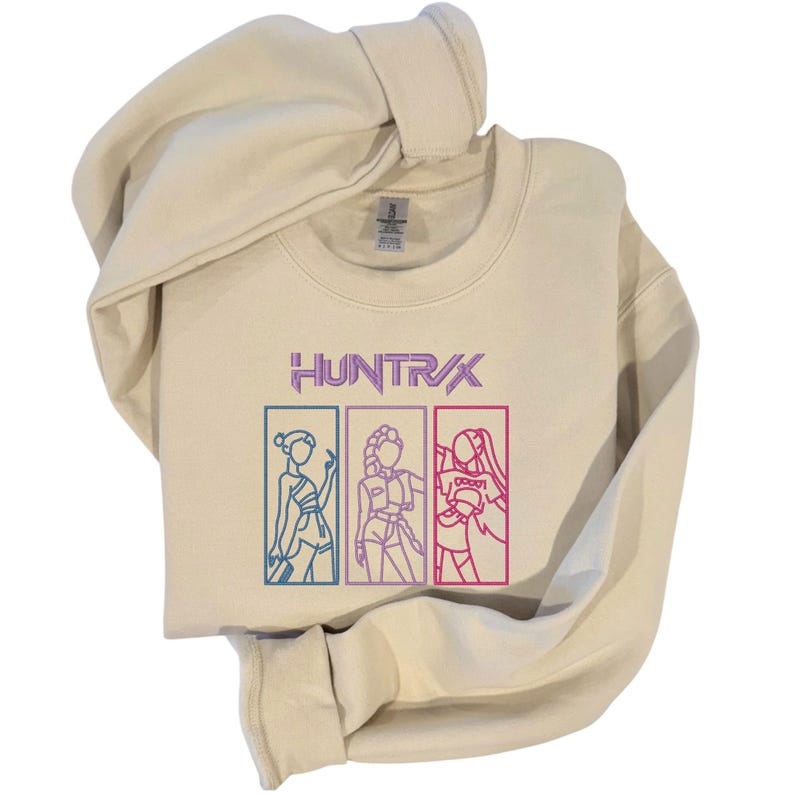 May include: Cream-colored sweatshirt with the word "HUNTR/X" in purple and three outlined figures in boxes. The figures are in blue, purple, and red. The sweatshirt is folded with the hood visible.