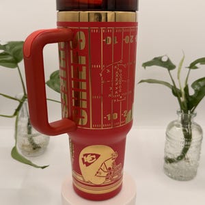 Puede incluir: Un vaso t&eacute;rmico rojo y dorado con asa. El vaso presenta un dise&ntilde;o de campo de f&uacute;tbol americano y el logotipo de los Kansas City Chiefs. La tapa y el asa son rojas. Dise&ntilde;ado para bebidas fr&iacute;as o calientes.