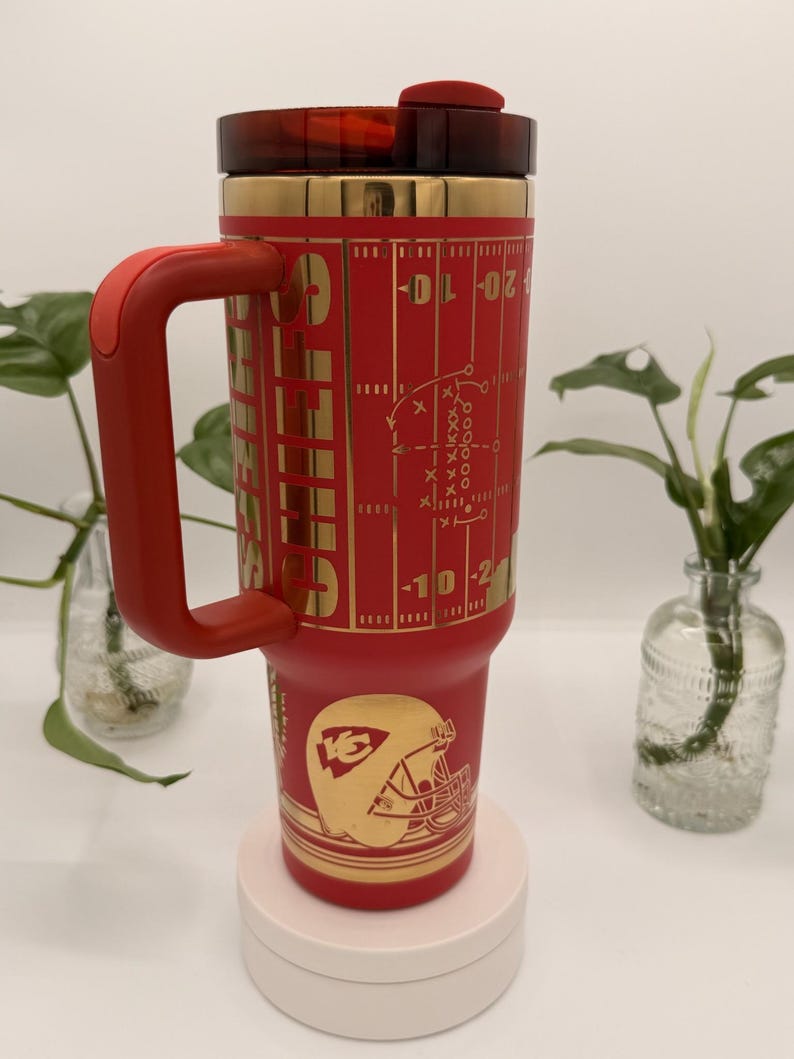 Puede incluir: Un vaso t&eacute;rmico rojo y dorado con asa. El vaso presenta un dise&ntilde;o de campo de f&uacute;tbol americano y el logotipo de los Kansas City Chiefs. La tapa y el asa son rojas. Dise&ntilde;ado para bebidas fr&iacute;as o calientes.
