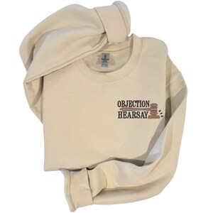 May include: Cream-colored sweatshirt with the words "Objection Hearsay" and a gavel graphic in brown. The sweatshirt has a crew neck and long sleeves. The fabric appears soft and comfortable.