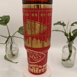 Puede incluir: Un vaso t&eacute;rmico rojo y dorado con el logotipo de los Kansas City Chiefs y marcas de campo de f&uacute;tbol. El vaso tiene una tapa roja y el texto "SUNDAYS ARE FOR THE CHIEFS".