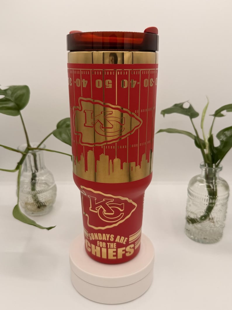 Puede incluir: Un vaso t&eacute;rmico rojo y dorado con el logotipo de los Kansas City Chiefs y marcas de campo de f&uacute;tbol. El vaso tiene una tapa roja y el texto "SUNDAYS ARE FOR THE CHIEFS".