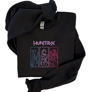 May include: Black sweatshirt with the word "HUNTR/X" in purple and three outlined figures in blue and pink. The sweatshirt has a crew neck and a hood.