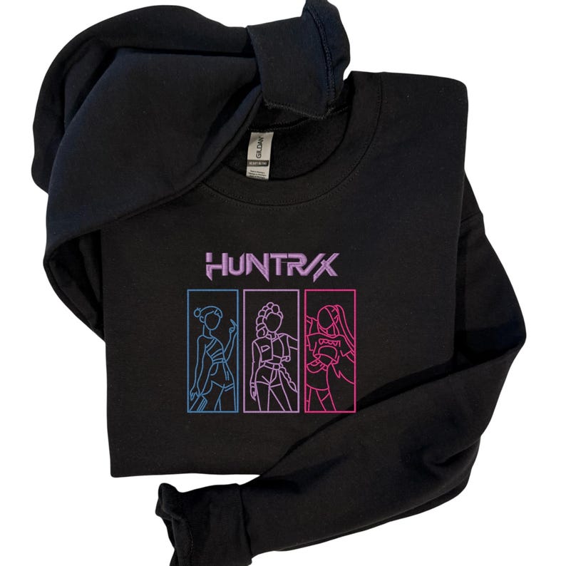 May include: Black sweatshirt with the word "HUNTR/X" in purple and three outlined figures in blue and pink. The sweatshirt has a crew neck and a hood.