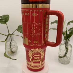 Puede incluir: Un termo de viaje rojo y dorado con asa. El termo presenta un dise&ntilde;o de campo de f&uacute;tbol y las palabras "Chiefs Kingdom" con un gr&aacute;fico de tocado nativo americano. La tapa y el asa son rojas.