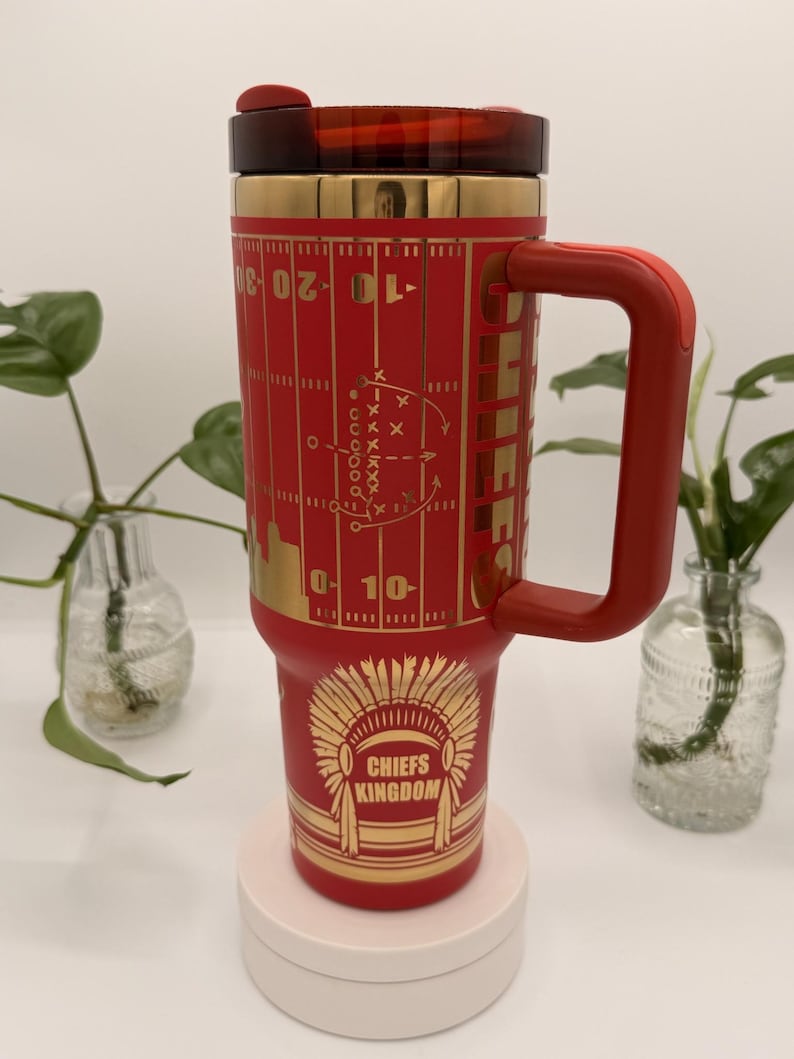 Puede incluir: Un termo de viaje rojo y dorado con asa. El termo presenta un dise&ntilde;o de campo de f&uacute;tbol y las palabras "Chiefs Kingdom" con un gr&aacute;fico de tocado nativo americano. La tapa y el asa son rojas.