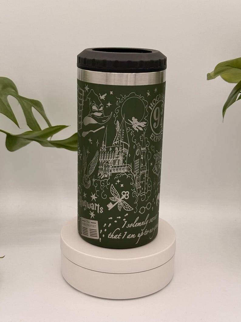 May include: Green insulated drinks holder with a black top and silver rim. The holder features white line art of Hogwarts, a key, and the text "I solemnly swear that I am up to no good." The holder is on a white stand.