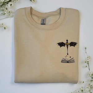 May include: A beige sweatshirt with a black embroidered dragon flying above an open book with stars. The book and dragon are embroidered in black with gold accents.