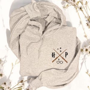May include: A gray hooded sweatshirt with a black embroidered design featuring crossed wands, the letters "H" and "P", and a pair of glasses.
