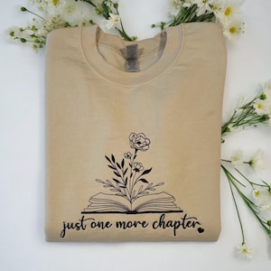 May include: A beige sweatshirt with a black graphic of an open book with flowers growing out of it. The text "just one more chapter" is written below the book.