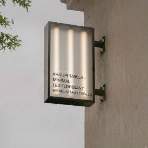 May include: Rectangular, black-framed sign mounted on a light-colored wall. The sign features three vertical, illuminated light bars behind a frosted panel. Text on the sign reads "KANOPİ TABELA - MİNİMAL LED FLORESAN AYDINLATMALI TABELA."