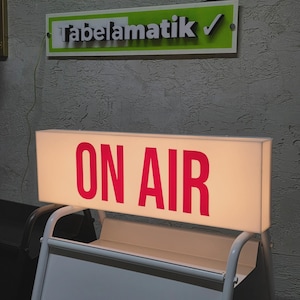 Peut inclure: Un panneau lumineux rectangulaire avec l'inscription "ON AIR" en lettres rouges. Il est monté sur un support en métal blanc. Au-dessus, un panneau indique "Tabelamatik" en lettres noires avec un bord vert et blanc. Le fond est un mur gris texturé.