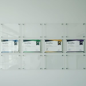 May include: A wall display featuring clear acrylic frames holding certificates. Four certificates are visible, each with a different colored border and the word "Sertifika" printed on them. The frames are mounted with silver hardware.