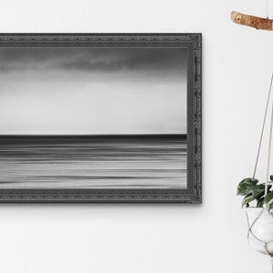 May include: A black and white photograph of a calm ocean with a cloudy sky. The photo is framed in a black ornate frame.