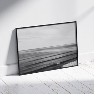 May include: A black and white photograph of ocean waves crashing on the shore. The photo is framed in a black frame and is leaning against a white wall.