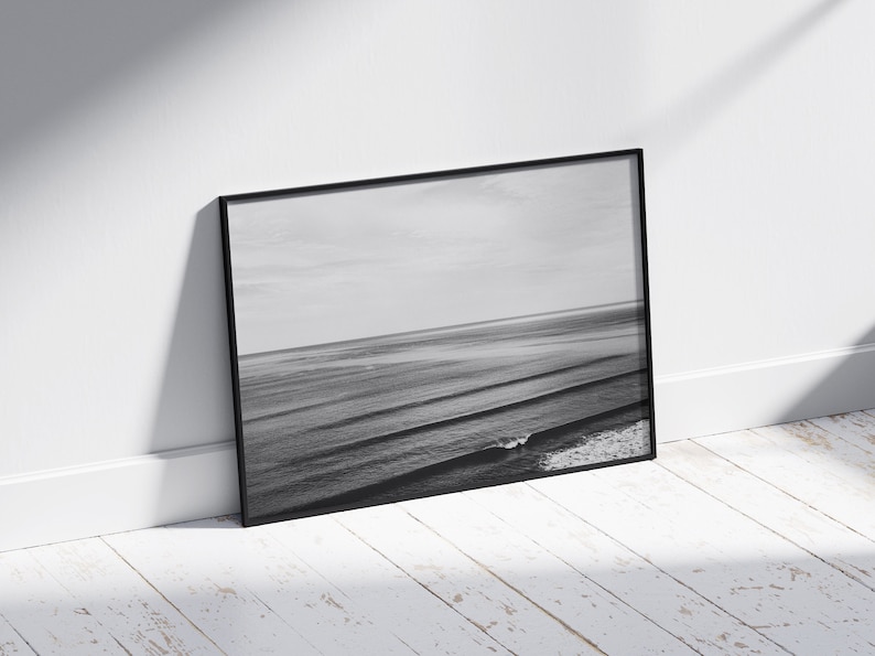 May include: A black and white photograph of ocean waves crashing on the shore. The photo is framed in a black frame and is leaning against a white wall.