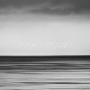 May include: A black and white photograph of a calm ocean with a cloudy sky. The text "Rhiain St. Aubo" is written in white across the middle of the image.