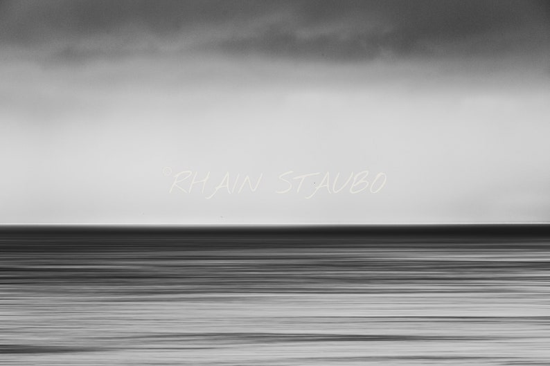 May include: A black and white photograph of a calm ocean with a cloudy sky. The text "Rhiain St. Aubo" is written in white across the middle of the image.
