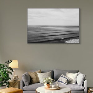 May include: A black and white photograph of ocean waves crashing on the shore. The photo is framed and hanging on a gray wall above a gray couch with pillows and a coffee table.