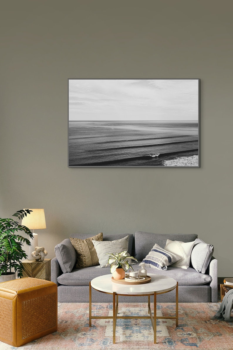 May include: A black and white photograph of ocean waves crashing on the shore. The photo is framed and hanging on a gray wall above a gray couch with pillows and a coffee table.
