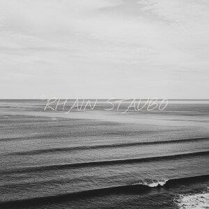 May include: A black and white photograph of a calm ocean with rolling waves. The horizon is in the distance and the sky is cloudy. The text "Rhiain Staubo" is written in white across the middle of the image.