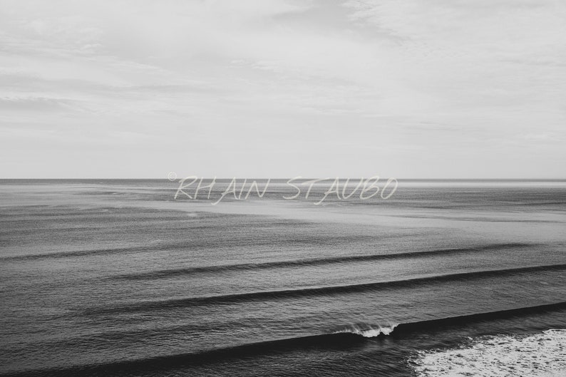 May include: A black and white photograph of a calm ocean with rolling waves. The horizon is in the distance and the sky is cloudy. The text "Rhiain Staubo" is written in white across the middle of the image.