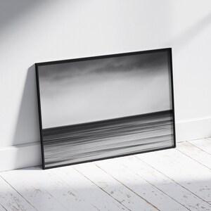 May include: A black and white photograph of a seascape with a cloudy sky and a horizon line. The photo is framed in a black frame and is leaning against a white wall.