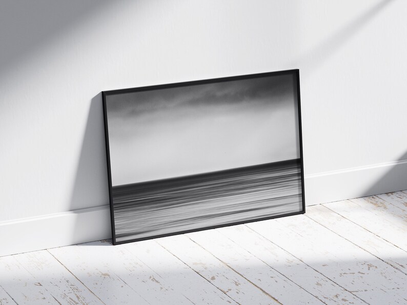 May include: A black and white photograph of a seascape with a cloudy sky and a horizon line. The photo is framed in a black frame and is leaning against a white wall.