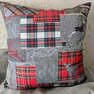 May include: Square patchwork cushion with a variety of fabrics in red, grey, and white. The cushion features plaid patterns, knitted textures, and a deer silhouette. A decorative home accent.