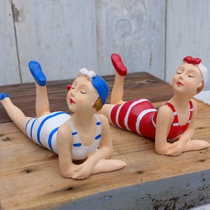 May include: Two ceramic figurines of women wearing vintage bathing suits. The woman on the left is wearing a blue and white striped swimsuit and the woman on the right is wearing a red and white striped swimsuit. Both women are lying on their stomachs with their heads turned to the side.