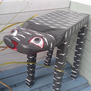 May include: A wooden cat-shaped stool with black and white stripes painted on the top and legs. The stool has a red nose and large eyes.