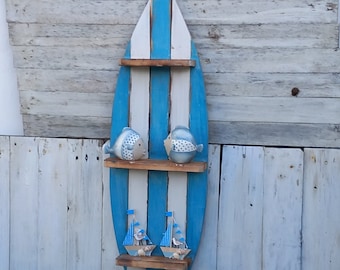 May include: A surfboard-shaped wall shelf painted in blue and white stripes. Two wooden shelves hold ceramic fish and sailboat decorations. The shelf is mounted on a weathered, light-colored wooden wall.