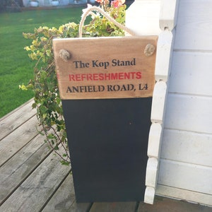May include: A wooden sign with a black chalkboard hanging on a white wooden wall. The sign says "The Kop Stand Refreshments Anfield Road, L4".
