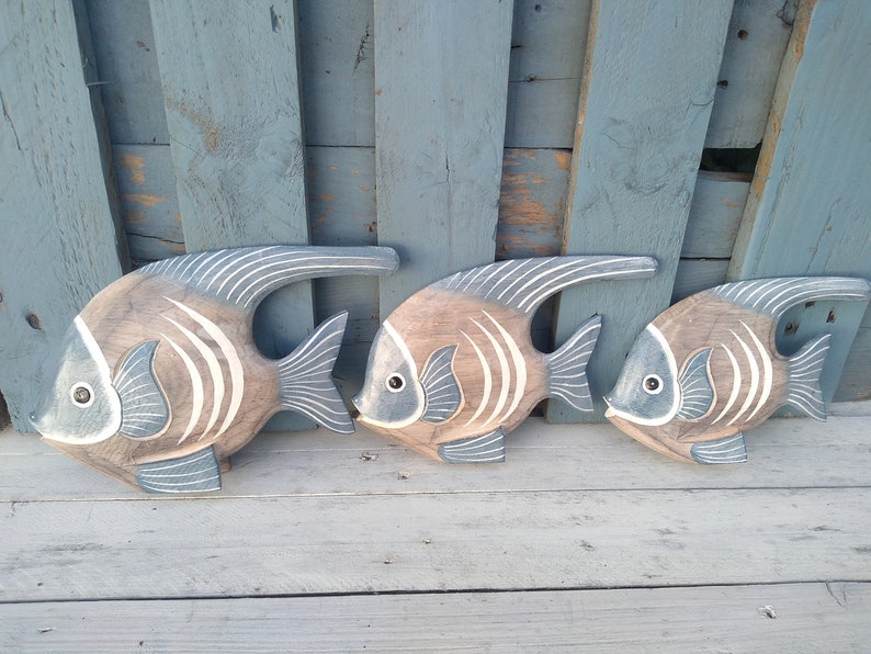 May include: Three wooden fish wall decor in shades of brown and blue with white stripes. The fish are arranged in a row, with the largest fish on the left and the smallest fish on the right.