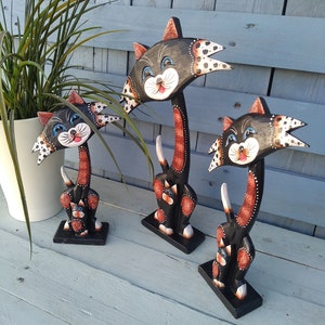 Peut inclure: Trois figurines de chat en bois peintes en noir, blanc et orange. Les chats sont debout sur une surface en bois bleu clair.
