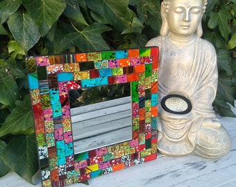 May include: A colorful mosaic mirror with a square frame. The frame is made of small, colorful tiles in shades of red, orange, yellow, green, blue, and purple. The mirror is reflecting a wooden surface. A small golden statue of Buddha is sitting on the wooden surface behind the mirror.