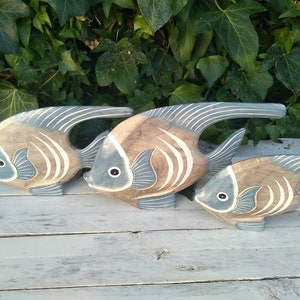 May include: Three wooden fish sculptures in shades of brown and blue, arranged in a row on a white wooden surface. The fish have a stylized design with white stripes and are facing the left.