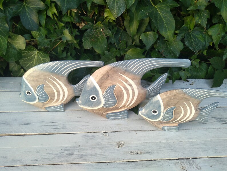 May include: Three wooden fish sculptures in shades of brown and blue, arranged in a row on a white wooden surface. The fish have a stylized design with white stripes and are facing the left.