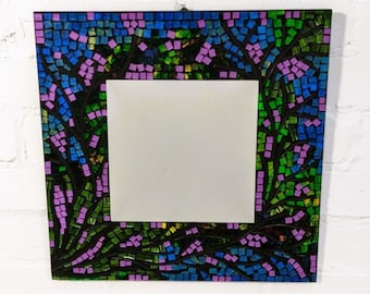 May include: A square mirror with a mosaic frame. The frame is made of small, colorful tiles in shades of green, blue, and purple. The tiles are arranged in a floral pattern.