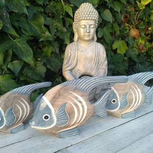 May include: Three wooden fish sculptures with blue and brown paint. The fish are arranged in a row on a white wooden surface. A golden Buddha statue is in the background.