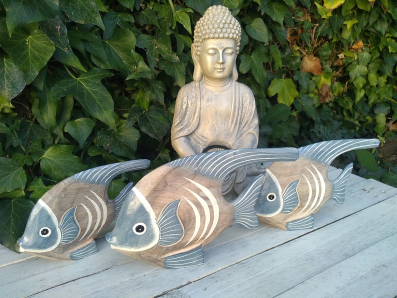May include: Three wooden fish sculptures with blue and brown paint. The fish are arranged in a row on a white wooden surface. A golden Buddha statue is in the background.