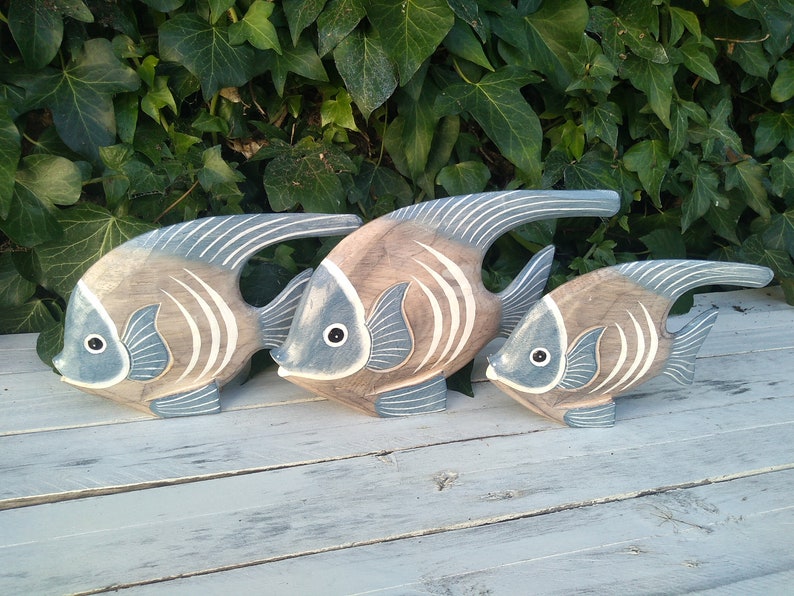 May include: Three wooden fish sculptures in shades of brown and blue, arranged in a row from largest to smallest. The fish have a stylized design with white stripes and are painted with a distressed finish.