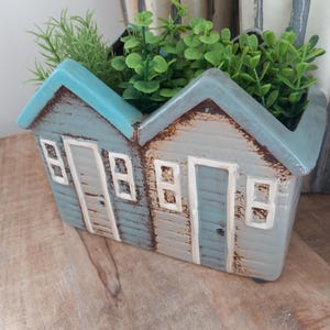 May include: A ceramic planter in the shape of two houses, painted in blue and grey with white detailing. The planter is filled with green artificial plants. It is placed on a wooden surface.