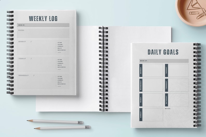 May include: Two spiral bound notebooks with gray covers. The left notebook has the text "WEEKLY LOG" and is divided into sections for each day of the week. The right notebook has the text "DAILY GOALS" and is divided into sections for each day of the week.
