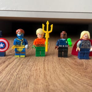 May include: Five Lego superhero minifigures are displayed on a wooden surface. The characters include Captain America with a shield, Cyclops, Aquaman with a trident, Nick Fury, and Thor with a hammer and cape. Each figure is brightly coloured.