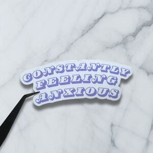 May include: A white sticker with the text "CONSTANTLY FEELING ANXIOUS" in a purple, retro font.
