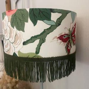 Puede incluir: Una pantalla de lámpara con un estampado floral que presenta flores rosas y beige, hojas verdes y una mariposa roja sobre un fondo blanco. El borde inferior está adornado con una franja verde oscuro.